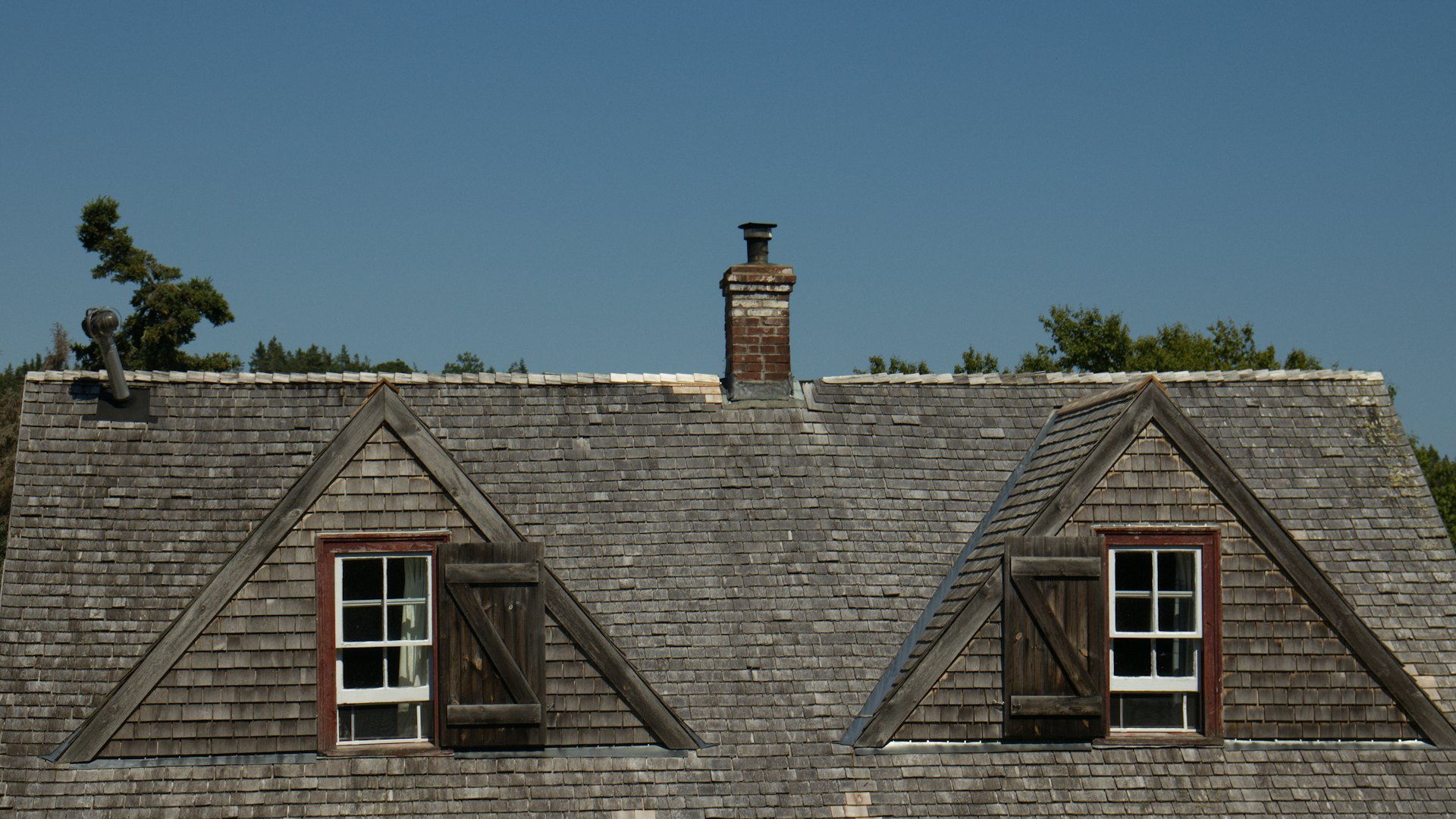 Two dormers on a weathered shingle roof with a brick chimney